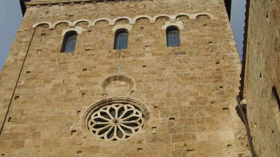Facciata in pietra di una chiesa storica vista dal basso, con rosone circolare scolpito, tre finestre ad arco e una fila di archetti decorativi sotto il timpano. In cima si trova un piccolo campanile a vela con campana, sullo sfondo di cielo nuvoloso.