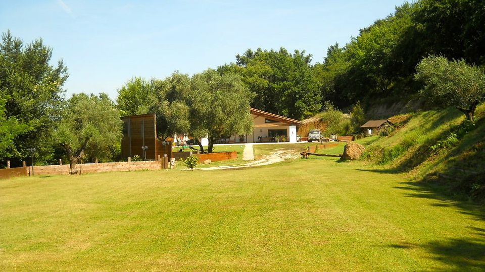 Ampio prato in una proprietà rurale con casetta a tetto spiovente, vialetto in ghiaia e piccole strutture in legno. Intorno ci sono alberi e un pendio boscoso che circondano l’area sotto un cielo sereno.