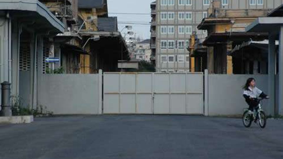 Strada asfaltata in un’area industriale con edifici bassi su entrambi i lati e un grande cancello metallico chiuso al centro. Sullo sfondo si vede un alto edificio residenziale con molte finestre e cavi sospesi sopra la carreggiata.