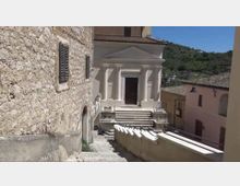 Scalinata in pietra tra edifici in muratura che conduce a una chiesa con facciata chiara, frontone triangolare e portico con colonne. Sullo sfondo si vedono case del borgo e colline verdi sotto un cielo sereno.