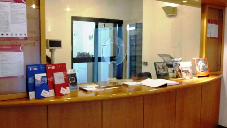 Banco reception in legno con ampia vetrata e sportello di servizio, con opuscoli e documenti esposti sul piano. Sullo sfondo si vede un corridoio con porta vetrata e pareti chiare illuminate da faretti a soffitto.