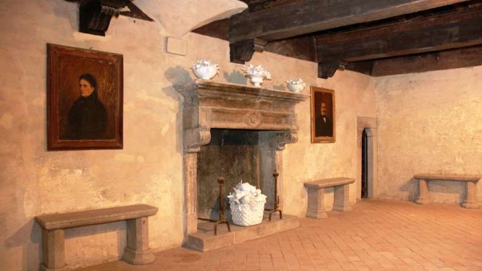 Interno di una sala storica con grande camino in pietra al centro, soffitto con travi di legno a vista e pavimento in cotto. Alle pareti intonacate sono appesi ritratti incorniciati e sono presenti panche in pietra lungo i lati.
