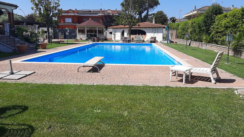 Piscina rettangolare all’aperto con bordo piastrellato e acqua azzurra, circondata da prato curato e lettini. Sullo sfondo si vedono un gazebo e una piccola struttura con portico ad archi e tetto in coppi, con palme e case residenziali intorno.