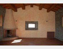 Interno rustico con pavimento in cotto e soffitto a travi di legno a vista, pareti intonacate e due pareti laterali in pietra. A sinistra c’è un grande camino ad angolo, al centro una piccola finestra con cornice in pietra e a destra un radiatore sotto applique a parete.