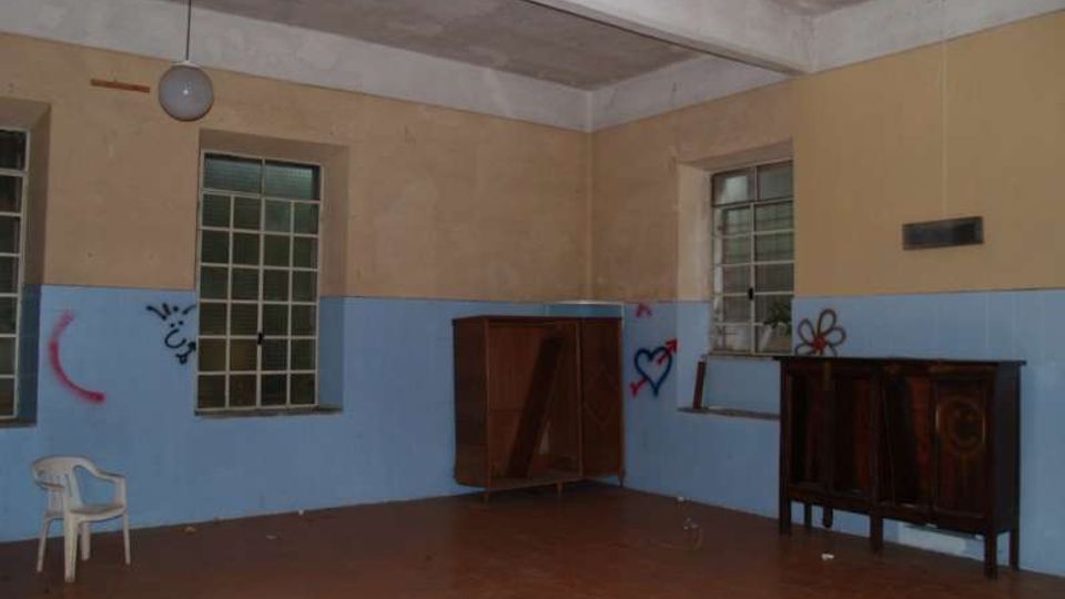 Interno di una stanza spoglia con pavimento in piastrelle marroni, pareti bicolori beige e azzurre e soffitto alto con lampada a sospensione. Due finestre con inferriate e alcuni mobili in legno lungo le pareti, con graffiti visibili sulle superfici.
