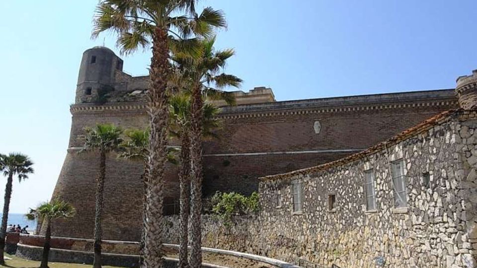 Fortezza costiera in mattoni con bastioni e torre angolare, affiancata da un lungo edificio in pietra con piccole finestre. In primo piano ci sono palme e un prato con percorso pedonale e ringhiera, sotto un cielo sereno.