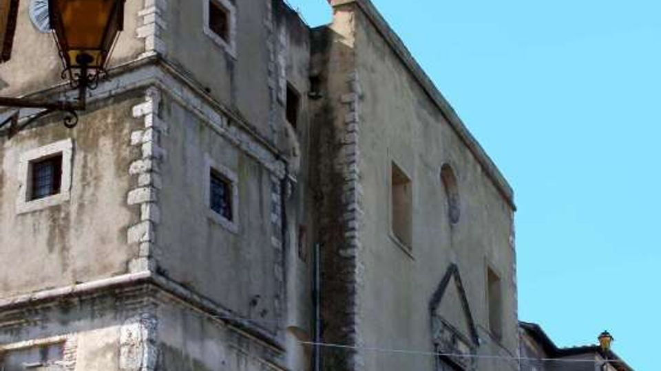 Chiesa in muratura intonacata con campanile a torre e piccole finestre, affacciata su una strada di paese. Una lunga scalinata in pietra conduce all’ingresso laterale, con lampioni e alcune auto parcheggiate in primo piano.