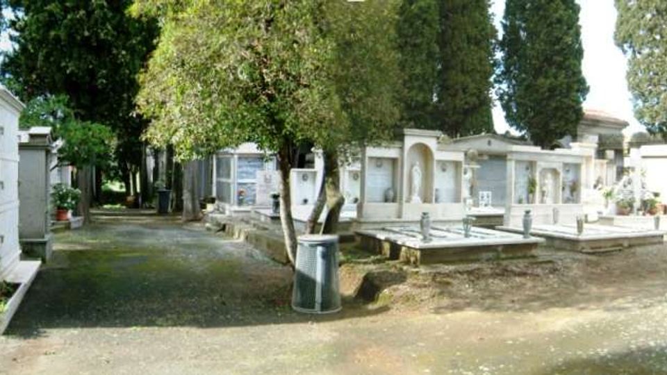 Viale di un cimitero con vialetto in ghiaia affiancato da tombe e mausolei in pietra e marmo, con piccole cappelle e lapidi. Alberi alti e cipressi circondano l’area, con una panchina e un cestino lungo il percorso.