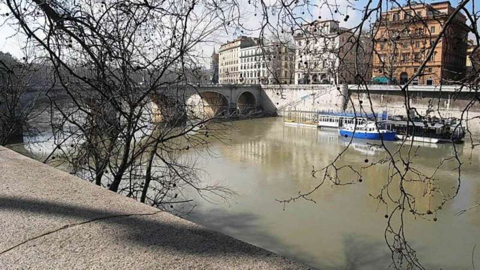 Veduta di un fiume cittadino con un ponte in pietra a più arcate, edifici storici lungo la sponda e alcune barche ormeggiate. In primo piano si vedono rami spogli di alberi e un parapetto in pietra del lungofiume.