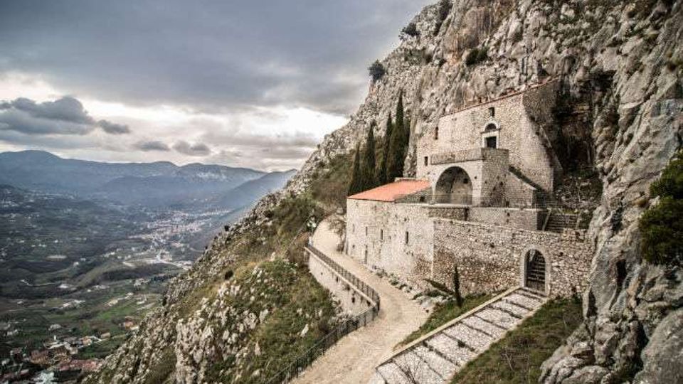 Santuario in pietra addossato a una parete rocciosa su un ripido versante montano, con terrazze murate, arco d’ingresso e sentiero curvo che costeggia l’edificio. Sullo sfondo si vede una valle ampia con colline e un cielo nuvoloso.