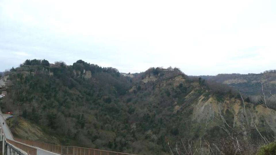 Veduta panoramica di una valle boscosa con colline e pareti di terreno eroso, osservata da un ponte o strada sopraelevata con parapetto metallico in primo piano. Sullo sfondo si vedono crinali e una scarpata chiara sotto un cielo coperto.