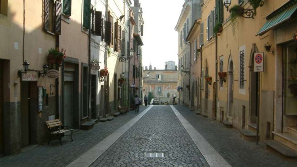 Strada stretta in pavé con marciapiedi in pietra, fiancheggiata da edifici storici a più piani con persiane, balconcini e lanterne a muro. Lungo le facciate si vedono ingressi di negozi, panchine e piante in vaso, con la strada che termina verso un edificio sullo sfondo.