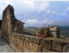 Ruderi in pietra di un edificio antico con muro basso e frammenti di muratura, affacciati su una valle collinare. Sullo sfondo si vedono campi e boschi sotto un cielo parzialmente nuvoloso.