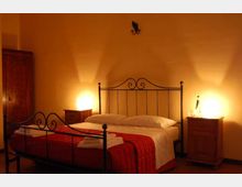 Camera da letto in stile tradizionale con letto matrimoniale in ferro battuto, copriletto rosso e biancheria piegata ai piedi. Due comodini in legno con lampade illuminano le pareti color crema, con armadio in legno sul lato sinistro.