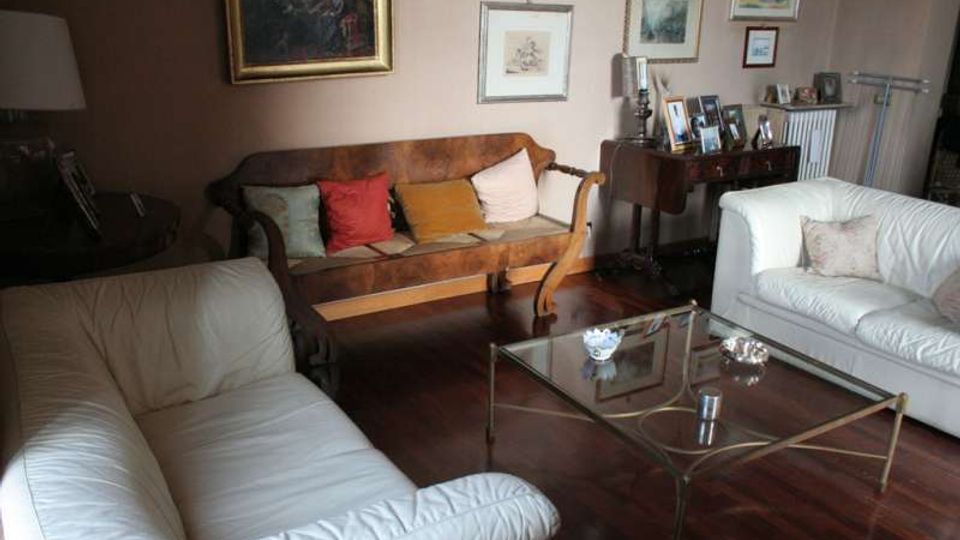 Soggiorno con pavimento in legno scuro, due divani bianchi e un divano in pelle marrone con cuscini colorati disposti attorno a un tavolino in vetro e metallo. Alle pareti sono appesi vari quadri e stampe, con una consolle con fotografie e una lampada vicino a un termosifone.