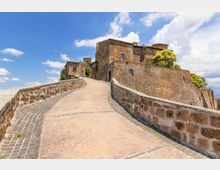 Rampa in pietra e mattoni con muretti laterali che conduce all’ingresso ad arco di un castello in pietra con alte mura e edificio fortificato. Sullo sfondo cielo azzurro con nuvole e vegetazione affacciata sulle mura.