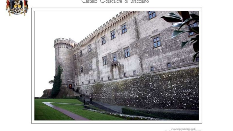 Veduta esterna del Castello Odescalchi di Bracciano, con alte mura in pietra, finestre regolari e una torre cilindrica merlata. In primo piano si vede un prato con un sentiero e il basamento del bastione lungo il perimetro del castello.