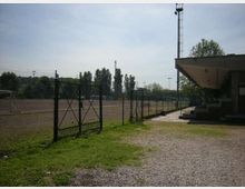 Campo sportivo all’aperto recintato con rete metallica e cancello, affiancato da un vialetto in ghiaia e da una piccola tribuna coperta sulla destra. Sullo sfondo si vedono alberi e alti pali per l’illuminazione sotto un cielo sereno.