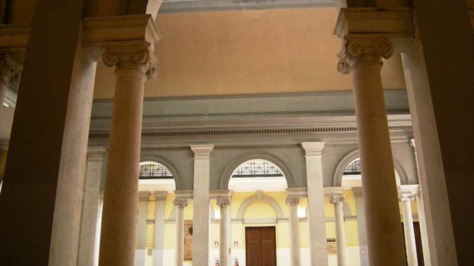 Cortile interno di un edificio storico con porticato ad archi e colonne in pietra con capitelli decorati. Sullo sfondo si vedono una facciata con finestre ad arco, una porta centrale in legno e un lucernario in alto.