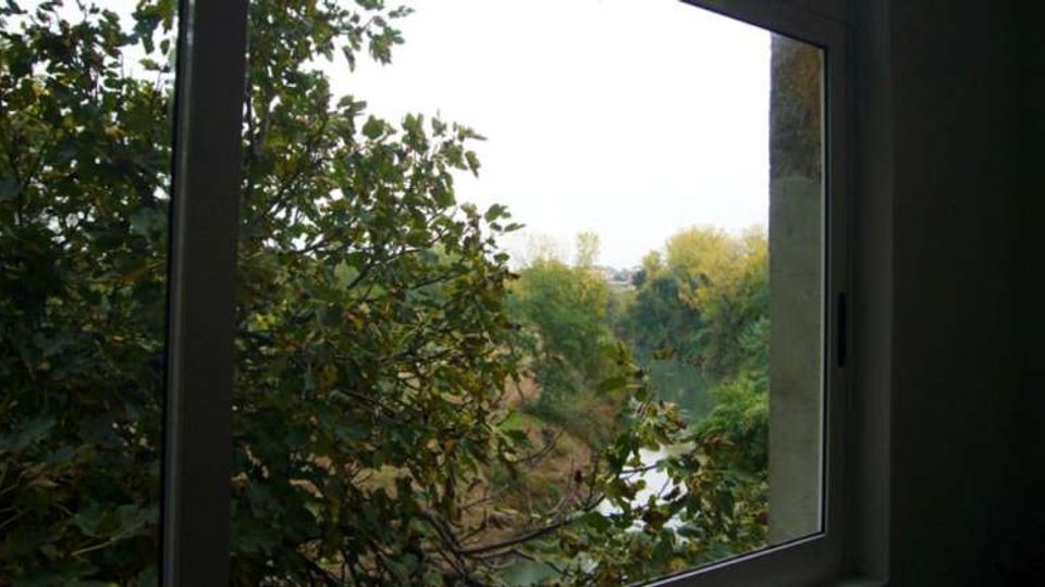 Vista dall’interno attraverso una finestra con telaio scuro su una zona verde con alberi e cespugli fitti. In lontananza si intravede un corso d’acqua che attraversa il paesaggio.