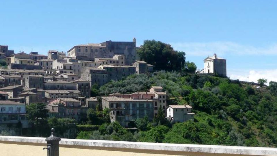 Veduta panoramica di un borgo collinare con case in pietra e tetti in coppi disposte a terrazze tra uliveti e vegetazione. Sulla cresta si distinguono un edificio storico e una piccola chiesa isolata sul pendio, sotto un cielo sereno.