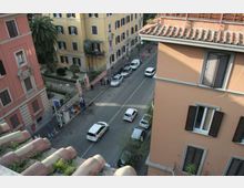 Vista dall’alto di una strada urbana con auto parcheggiate e in transito, fiancheggiata da edifici residenziali dai colori pastello con persiane e tetti in tegole. Sul lato sinistro si vede un piccolo giardino recintato con palme e un’area d’ingresso lungo il marciapiede.