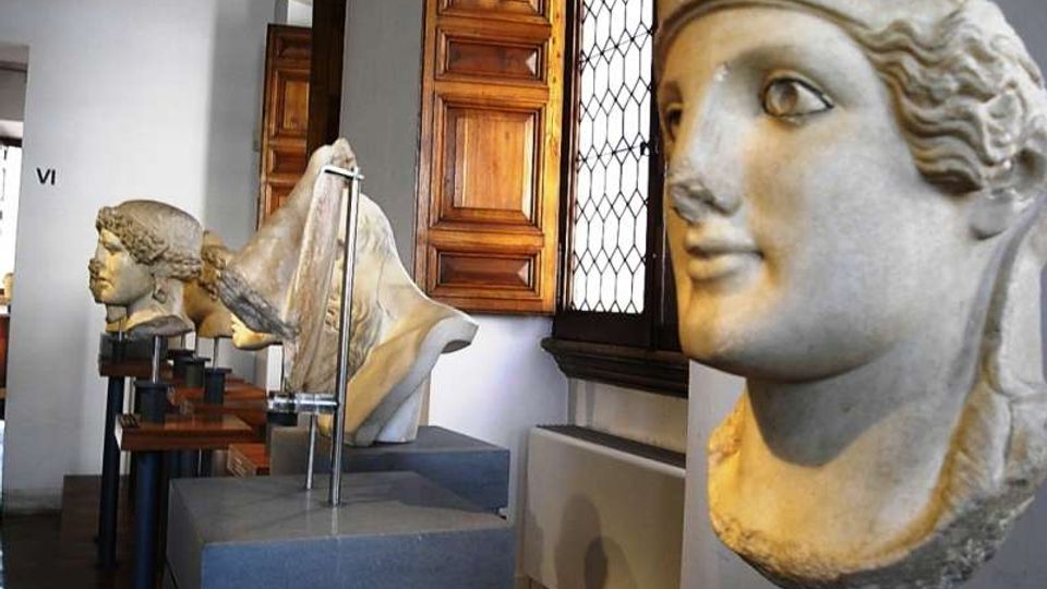 Interno di una sala museale con una fila di busti scultorei in pietra esposti su piedistalli lungo un corridoio. Sullo sfondo si vedono finestre con grata e persiane in legno, pareti chiare e pavimento in legno.