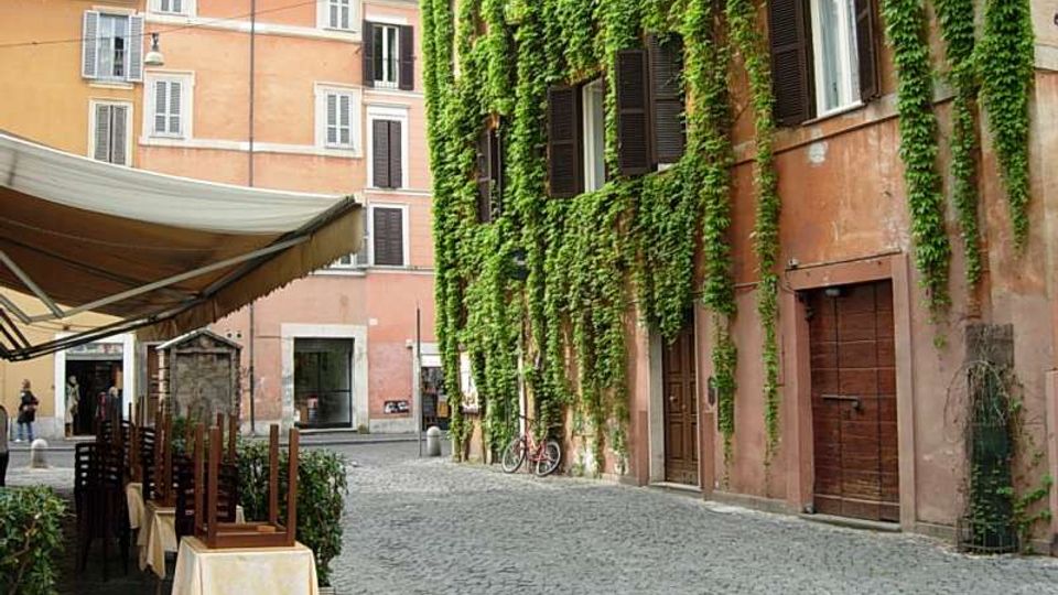 Strada acciottolata in una piazzetta urbana con palazzi dai toni rosa e arancioni; a destra un edificio con facciata coperta di edera e persiane scure. A sinistra si vede un’area esterna di ristorante con tavoli sotto una grande tenda e sedie impilate.