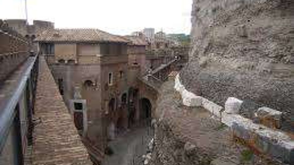 Vista da una passerella in pietra lungo una parete rocciosa, con parapetto e bandiera, che conduce verso un complesso di edifici storici in muratura e un arco su una strada sottostante. Sullo sfondo si vedono tetti in coppi e mura in pietra in un contesto di borgo antico.