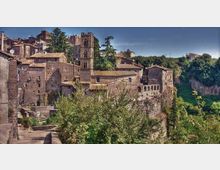 Borgo storico con edifici in pietra dai tetti in coppi, affacciato su una scarpata rocciosa sopra una valle boscosa. Al centro si distingue una torre campanaria e, lungo il bordo del dirupo, un edificio con loggiato ad archi.