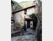 Casa in pietra addossata alla roccia lungo una stretta scalinata in selciato, con porta a griglia sotto una piccola tettoia in coppi, lanterna e piante rampicanti. Sullo sfondo si vedono montagne boscose e pareti rocciose.