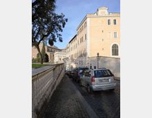 Strada in sampietrini in discesa fiancheggiata da un alto edificio storico intonacato color ocra con finestre regolari e da un muro in pietra con alberi sopra. Auto parcheggiate lungo il lato destro e, in fondo, un portale ad arco che conduce verso un complesso architettonico monumentale.