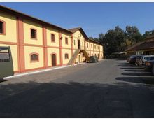 Edificio lungo a due piani in stile industriale, con facciata giallo chiaro e cornici rosse, finestre ad arco e scala esterna centrale che conduce a un ingresso sopraelevato. Davanti si estende un ampio piazzale asfaltato con auto parcheggiate sul lato destro e alberi sullo sfondo sotto un cielo sereno.