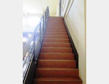 Scala interna in un edificio, con gradini in materiale rosso e corrimano metallico scuro sul lato sinistro. Pareti chiare e luce naturale proveniente dall’alto lungo il vano scala.