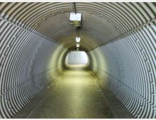 Sottopassaggio pedonale a forma di tunnel con pareti e soffitto in pannelli metallici ondulati, illuminato da lampade rettangolari a soffitto. Il pavimento asfaltato conduce verso un’uscita luminosa sul fondo.