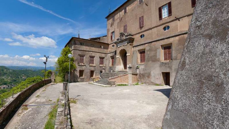 Cortile in cemento davanti a un grande edificio storico in pietra con finestre chiuse da persiane e un portale ad arco raggiungibile tramite una scalinata. Sul lato sinistro corre una strada con muretto e lampioni, affacciata su una valle collinare sotto cielo azzurro.