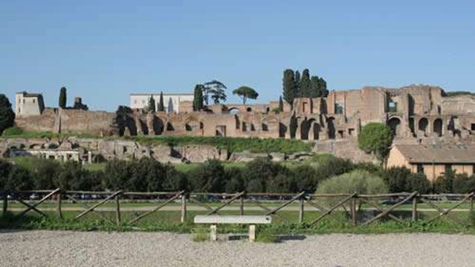 Veduta di un ampio complesso archeologico in muratura con resti di edifici e arcate su un pendio, con cipressi e altre alberature sullo sfondo. In primo piano si vede un’area verde delimitata da una staccionata e un sentiero ghiaioso con una panchina.