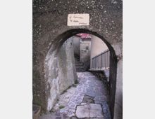 Arco in pietra scura che incornicia un vicolo stretto in discesa con pavimentazione irregolare in lastre di pietra, tra muri intonacati e una ringhiera metallica. In fondo si vedono scale, un tetto in coppi rossi e una targhetta con scritta sopra l’arco.