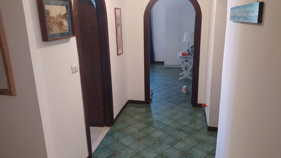 Corridoio interno di una casa con pareti bianche e pavimento in piastrelle verdi decorate, che conduce a due porte ad arco con cornici in legno scuro. Alle pareti sono appesi quadri e in fondo si intravedono un tavolino bianco con lampada e un armadio.