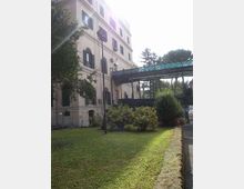 Edificio chiaro a più piani con finestre ad arco e persiane scure, affacciato su un prato con cespugli e alberi. A lato passa una passerella sopraelevata con parapetto verde, vicino a un vialetto asfaltato e a un lampione.