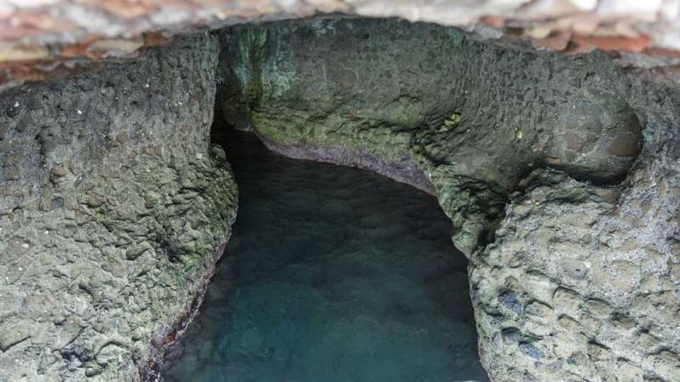 Apertura in muratura che affaccia su una cavità rocciosa con pareti irregolari e acqua limpida di colore turchese, simile a una piccola grotta o cisterna naturale. La vista è dall’alto, con i bordi in mattoni visibili lungo il perimetro.