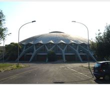 Edificio circolare con grande cupola ribassata e facciata scandita da archi e pilastri triangolari, con vetrate lungo il perimetro. Davanti si vede un ampio parcheggio asfaltato con lampioni e alberi ai lati.