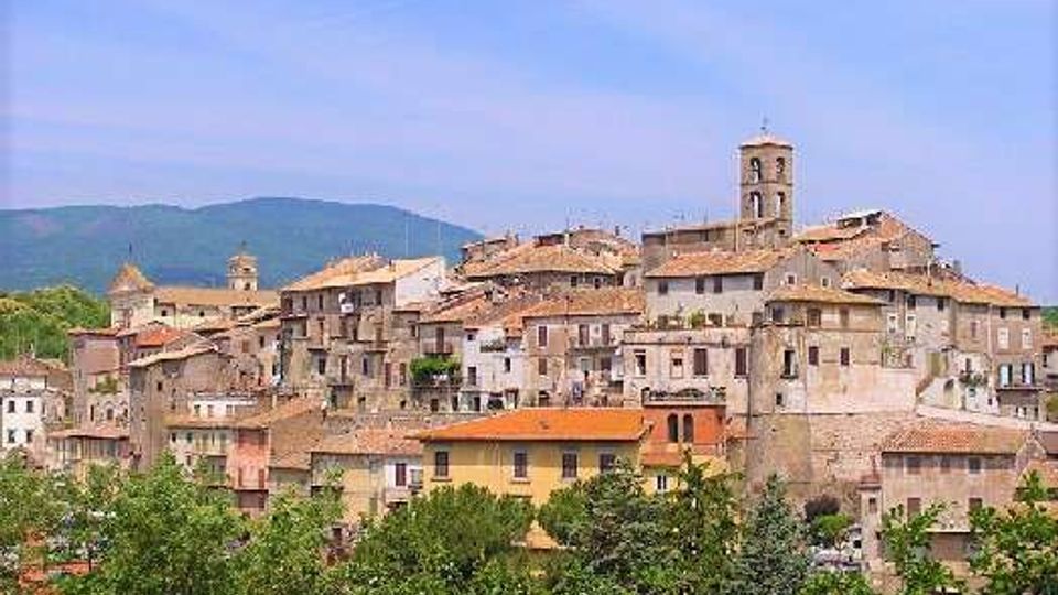 Vista panoramica di un borgo collinare con case in pietra addossate e tetti in coppi, dominato da un campanile e da edifici storici. In primo piano si vedono alberi e vegetazione, con colline sullo sfondo sotto un cielo sereno.