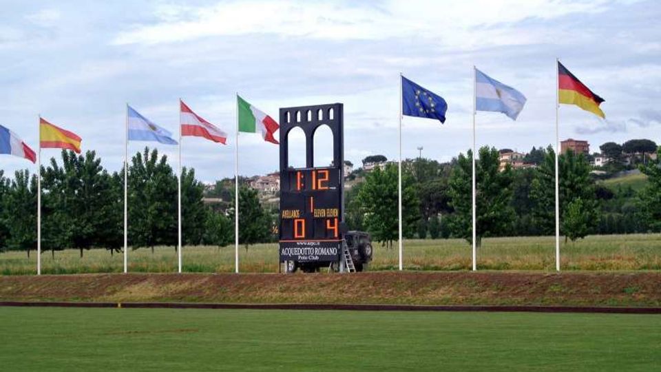 Tabellone segnapunti elettronico su un campo sportivo erboso, con una struttura centrale ad archi e cifre illuminate. Sullo sfondo una fila di alberi e diverse bandiere su alti pennoni in un’area verde collinare.