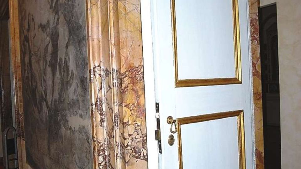 Interno di un edificio storico con pareti affrescate e rivestimenti in marmo venato; al centro una porta bianca con cornici dorate e maniglia in ottone. Pavimento in legno scuro e corridoio che prosegue sullo sfondo.