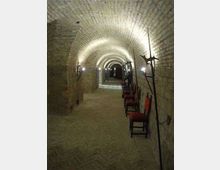Corridoio sotterraneo in pietra con soffitto a volta a botte, illuminato da lampade a parete. Lungo una parete sono disposte sedie e armi antiche esposte, con il passaggio che si estende fino a un’apertura in fondo.