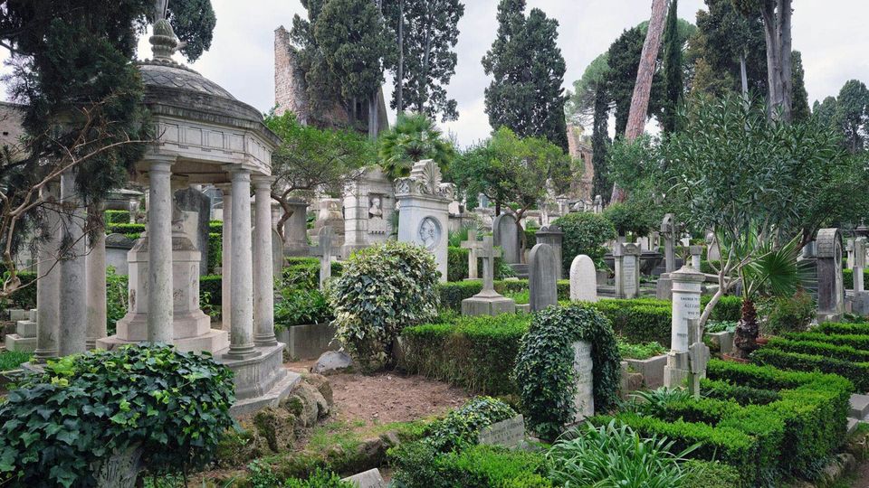 Veduta di un cimitero storico immerso nel verde, con numerose lapidi e monumenti in pietra tra siepi potate e vegetazione fitta. A sinistra si distingue un piccolo mausoleo circolare con colonne e cupola, circondato da alti cipressi e altri alberi.