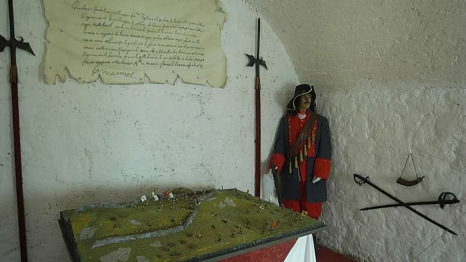 Piccola sala espositiva con pareti bianche in muratura, con una vetrina/maquette di terreno su un tavolo al centro e un cartiglio storico appeso alla parete. Alle pareti sono esposti attrezzi e armi antiche, tra cui una lunga alabarda e una bilancia, con una figura in uniforme d’epoca nell’angolo.