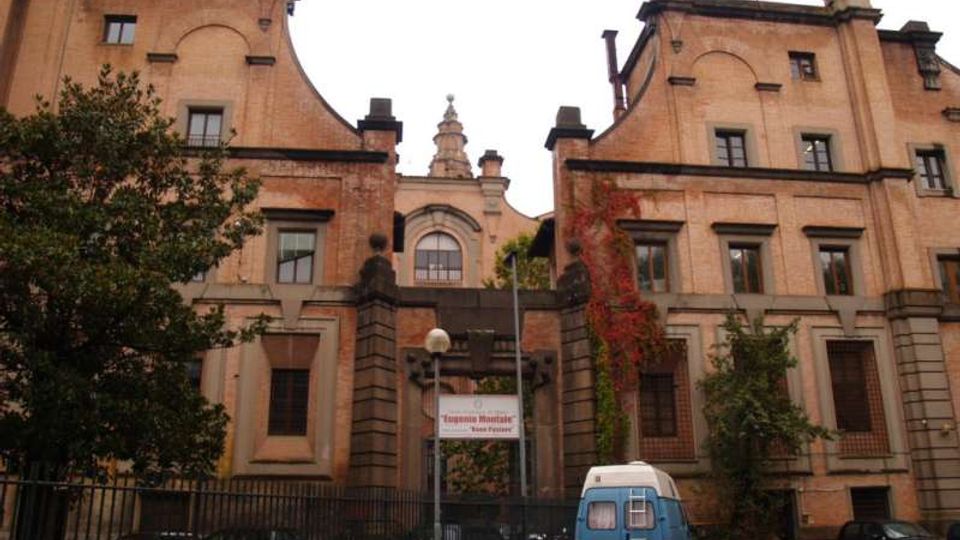 Facciata di un grande edificio storico in mattoni, simmetrico, con due corpi laterali e una parte centrale arretrata con frontone e finestrone ad arco. Davanti si vede un cancello in ferro, vegetazione rampicante e un’area di accesso con alcuni veicoli parcheggiati.