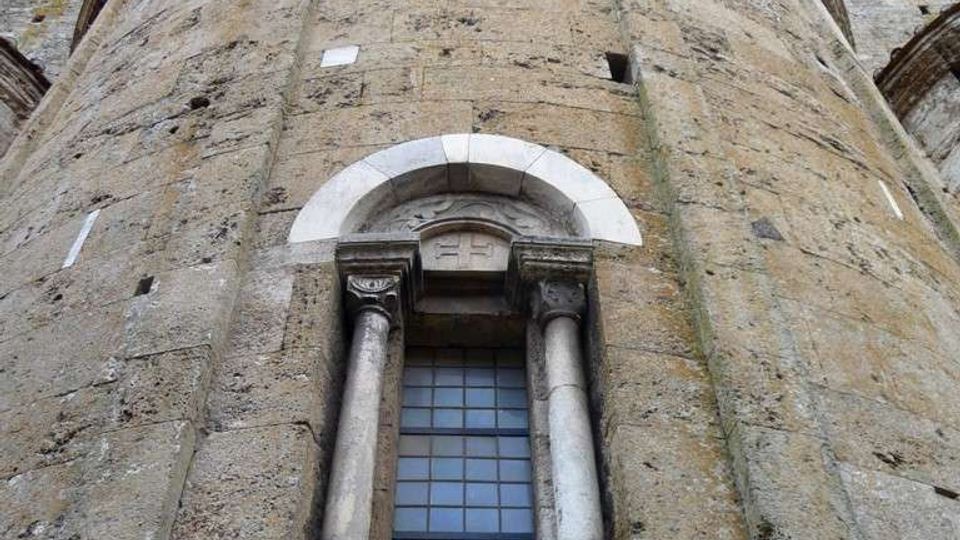 Vista dal basso di una torre cilindrica in pietra di un edificio storico, con piccola finestra ad arco incorniciata da due colonne e grata. In alto si nota un cornicione con archetti decorativi lungo il bordo.
